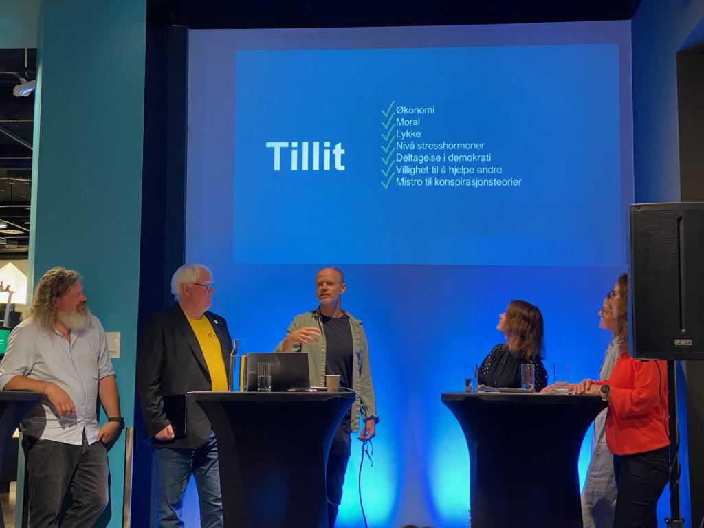 Torgeir Waterhouse, Terje André Olsen, Harald Eia, Kari Sand, Beth Stensen og Gunn Karin Gjul på scena i debatt under Arendalsuka. Tekst på storskjerm i bakgrunnen: "Tillit", med sjekklistepunktene: Økonomi, moral, lykke, nivå stresshormoner, deltagelse i demokrati, villighet til å hjelpe andre, mistro til konspirasjonsteorier.